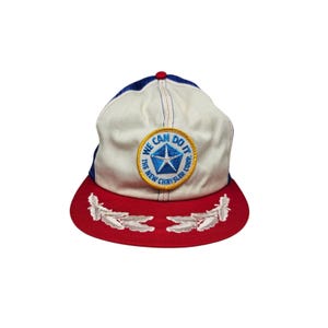 May include: A vintage baseball cap with a red peak and a white crown. The cap features a patch with the text "WE CAN DO IT" and "THE NEW CHRYSLER CORP" around a blue star logo. The cap has a blue and red colour scheme.
