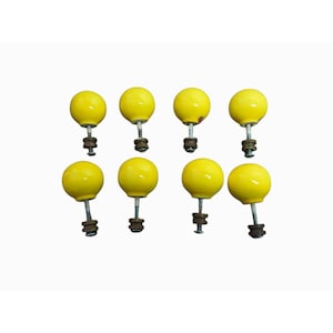 Puede incluir: Un juego de ocho pomos redondos para armarios de color amarillo brillante. Cada pomo tiene una cabeza esférica lisa unida a un perno metálico con una arandela y una tuerca oscuras en la base. Los pomos están dispuestos en dos filas de cuatro.