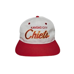 May include: A white and red baseball cap featuring the text "KANSAS CITY Chiefs" in gold and red lettering. The cap has a red peak and a red button. This is a sports-themed accessory.