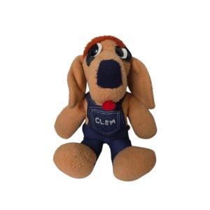 May include: A vintage plush dog toy with a brown body, floppy ears, and a red tongue. The dog wears blue denim overalls with the name "CLEM" on the bib. The toy has large, expressive eyes and a red hair-like patch.