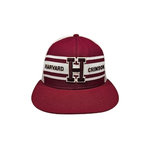 Puede incluir: Gorra de béisbol granate y blanca con el texto "HARVARD CRIMSON" en letras blancas. La letra "H" está en el centro. La gorra tiene una visera curva y una parte trasera de malla.