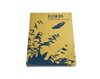 May include: A hardcover book titled "Illusions" by Richard Bach. The cover is gold with blue and black abstract designs and text. A feather graphic is also on the cover. The book is in good condition.
