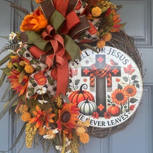 May include: A fall-themed wreath with a wooden base, adorned with orange sunflowers, pumpkins, and decorative ribbons. The wreath features a central circular sign with a cross, pumpkins, and the text "FALL FOR JESUS HE NEVER LEAVES."