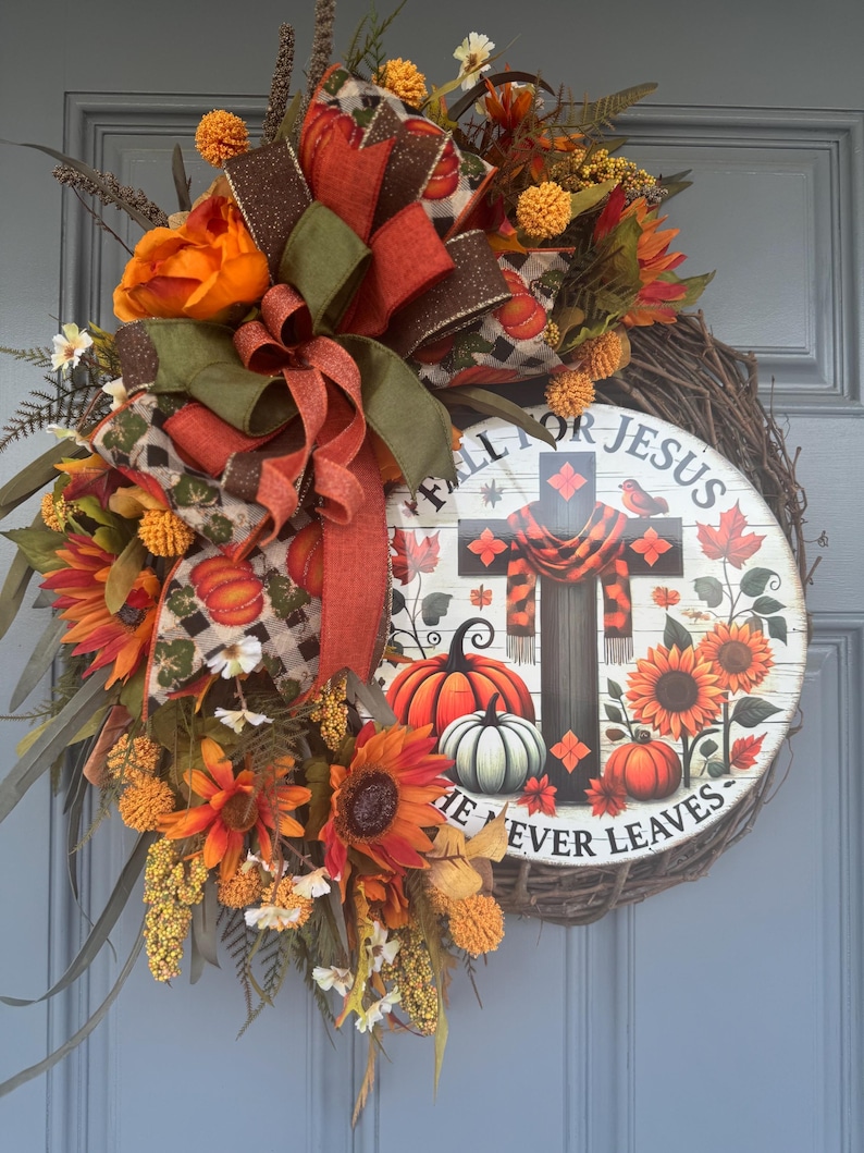 May include: A fall-themed wreath with a wooden base, adorned with orange sunflowers, pumpkins, and decorative ribbons. The wreath features a central circular sign with a cross, pumpkins, and the text "FALL FOR JESUS HE NEVER LEAVES."