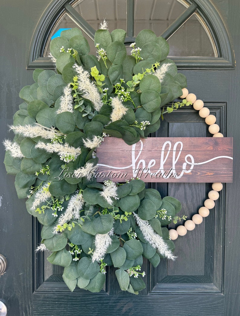May include: A green eucalyptus and white pampas grass wreath with a wooden sign that reads 'hello'. The wreath is decorated with wooden beads.