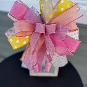 Peut inclure: Un nœud en ruban rose, jaune et blanc à pois avec un ruban pailleté rose. Le nœud est noué autour d'une lanterne blanche.