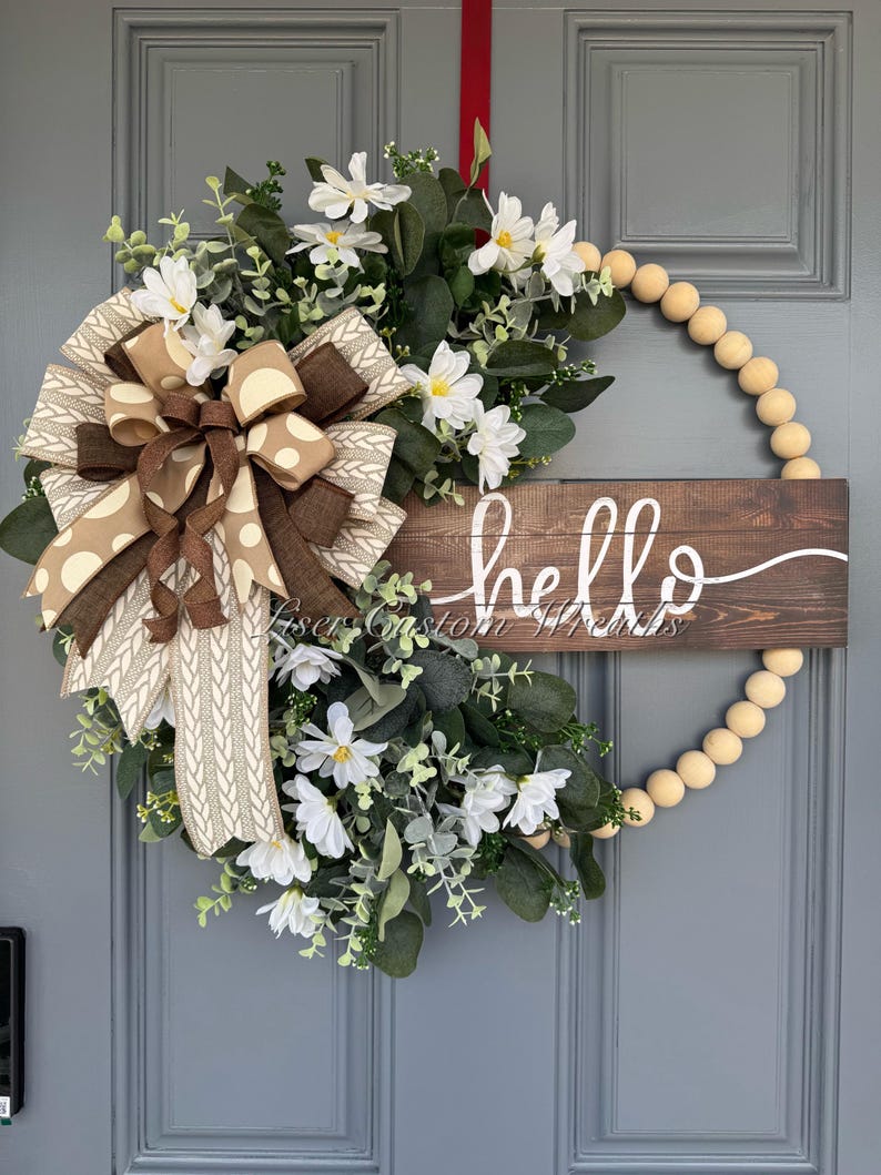 May include: A wooden beaded wreath with a brown and white polka dot bow and white flowers. A wooden sign with the word 'hello' is attached to the wreath.