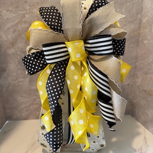 May include: A decorative bow with a bee theme. The bow uses ribbons in black and white polka dots, black and white stripes, and yellow with white polka dots. It also includes a beige burlap ribbon and a ribbon with a bee pattern.