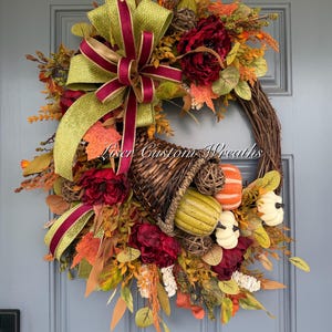 May include: A decorative wreath featuring a woven grapevine base, a large green and burgundy ribbon, dark red flowers, and a central cornucopia filled with small pumpkins and gourds in orange, green, and white. The text Liser Custom Wreaths is visible.
