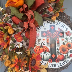 May include: A fall-themed wreath with a circular sign that reads "Fall for Jesus, He Never Leaves." The wreath features sunflowers, pumpkins, and a cross design. The wreath is decorated with orange, green, and brown ribbons and flowers.