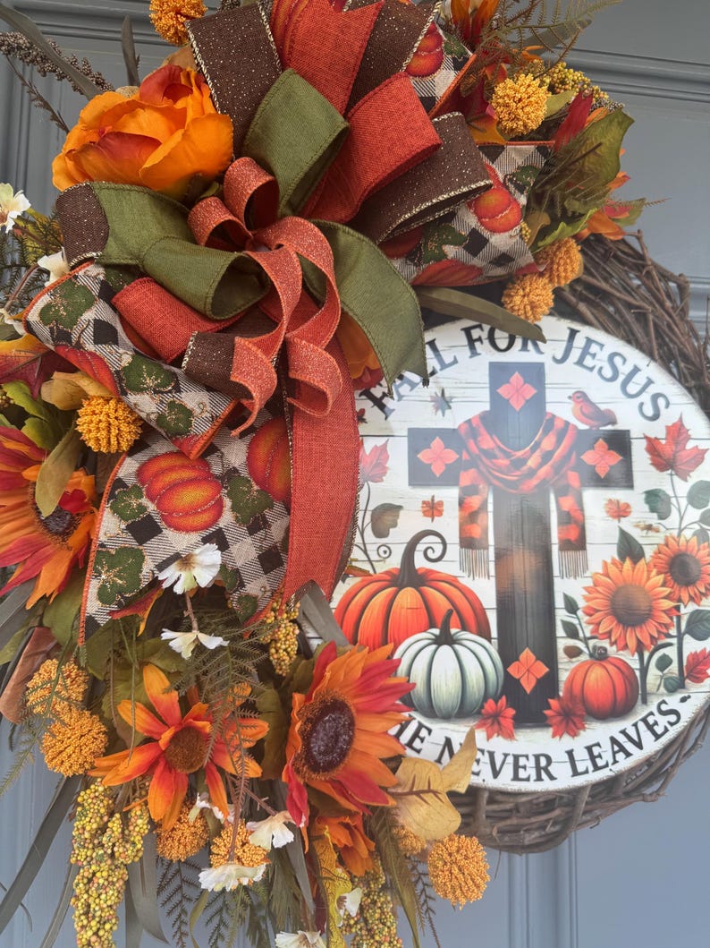 May include: A fall-themed wreath with a circular sign that reads "Fall for Jesus, He Never Leaves." The wreath features sunflowers, pumpkins, and a cross design. The wreath is decorated with orange, green, and brown ribbons and flowers.