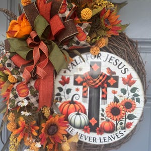 May include: A fall-themed wreath with sunflowers, pumpkins, and a decorative sign that reads "Fall for Jesus, He Never Leaves." The wreath features a mix of orange, brown, and green ribbons and faux foliage. The sign has a cross design with a scarf.