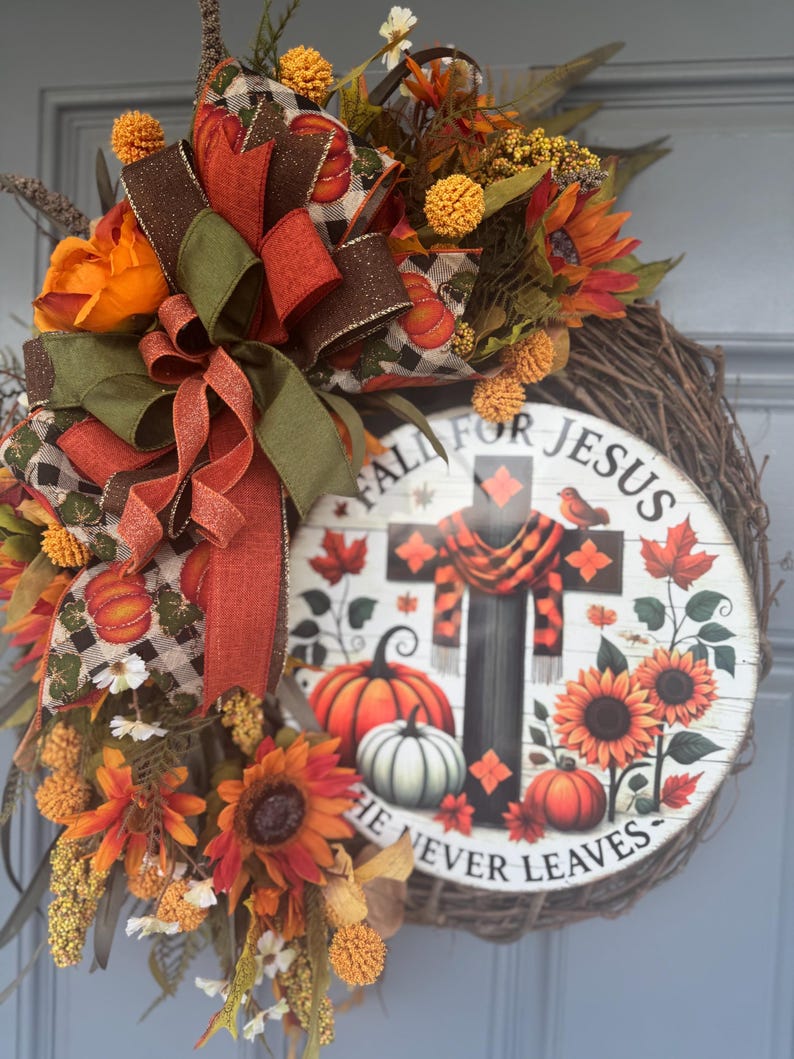 May include: A fall-themed wreath with sunflowers, pumpkins, and a decorative sign that reads "Fall for Jesus, He Never Leaves." The wreath features a mix of orange, brown, and green ribbons and faux foliage. The sign has a cross design with a scarf.