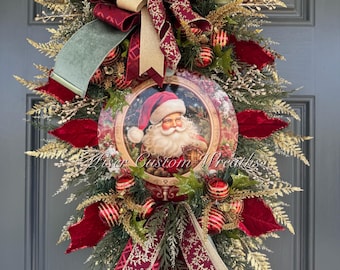May include: A Christmas door swag with a red velvet bow, gold and red ribbon, and a round ornament featuring a vintage illustration of Santa Claus. The swag is decorated with greenery, red and gold ornaments, and a green velvet ribbon.