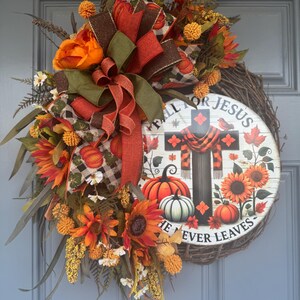 May include: A fall-themed wreath with a wooden sign that reads "FALL FOR JESUS HE NEVER LEAVES." The wreath features sunflowers, pumpkins, and a cross design. It is decorated with orange, brown, and green ribbons and foliage.