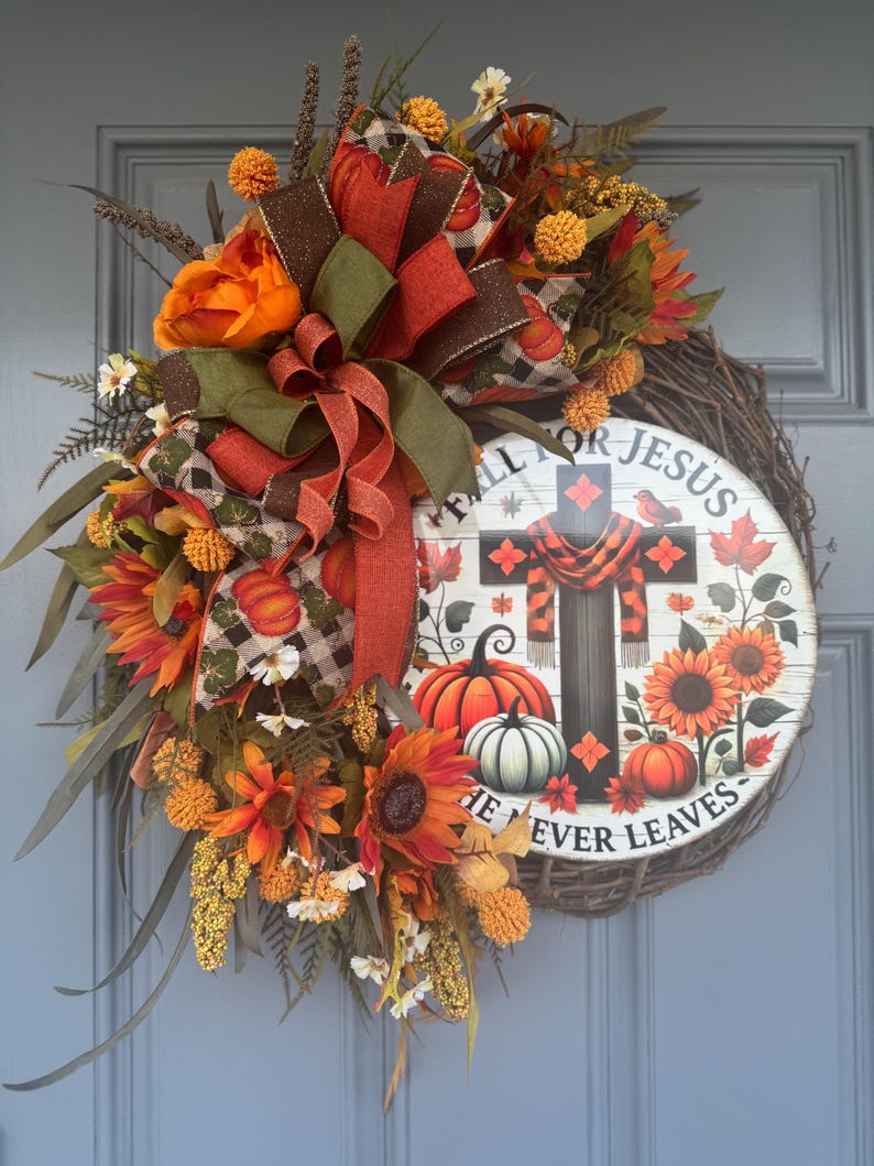 May include: A fall-themed wreath with a wooden sign that reads "FALL FOR JESUS HE NEVER LEAVES." The wreath features sunflowers, pumpkins, and a cross design. It is decorated with orange, brown, and green ribbons and foliage.