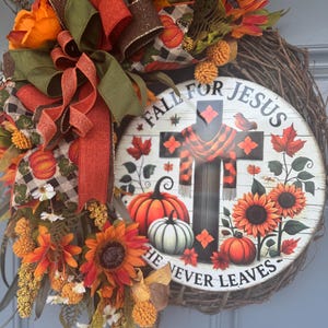 May include: A fall-themed wreath with a rustic grapevine base. It features sunflowers, pumpkins, and a sign that reads "Fall for Jesus, He Never Leaves." The wreath is adorned with orange, brown, and green ribbons and decorative accents.