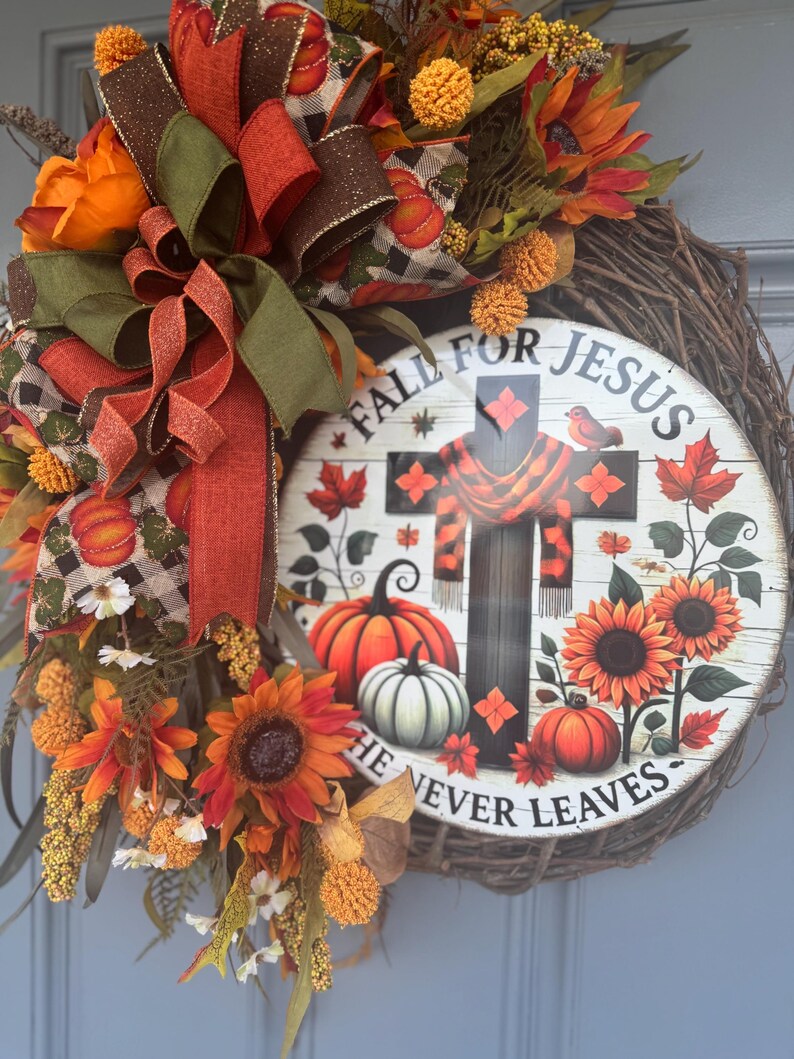 May include: A fall-themed wreath with a rustic grapevine base. It features sunflowers, pumpkins, and a sign that reads "Fall for Jesus, He Never Leaves." The wreath is adorned with orange, brown, and green ribbons and decorative accents.