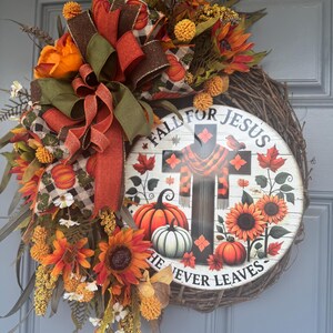 May include: A fall-themed wreath with a wooden base, adorned with sunflowers, pumpkins, and decorative ribbons in orange, brown, and green. The wreath features a circular sign with the text "Fall for Jesus, He Never Leaves."