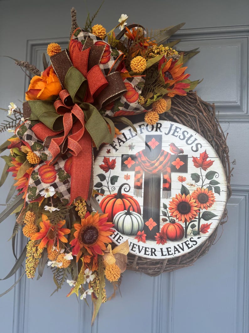 May include: A fall-themed wreath with a wooden base, adorned with sunflowers, pumpkins, and decorative ribbons in orange, brown, and green. The wreath features a circular sign with the text "Fall for Jesus, He Never Leaves."