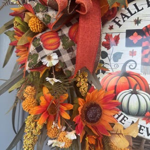 May include: A fall-themed wreath with sunflowers, pumpkins, and fall foliage. The wreath features a wooden sign with the words "Fall Fun" and a pumpkin illustration. The wreath is decorated with orange, red, and green ribbons and accents.
