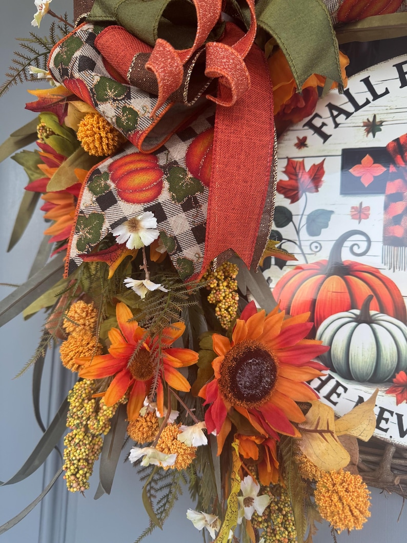 May include: A fall-themed wreath with sunflowers, pumpkins, and fall foliage. The wreath features a wooden sign with the words "Fall Fun" and a pumpkin illustration. The wreath is decorated with orange, red, and green ribbons and accents.