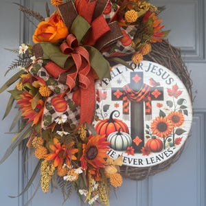 May include: A fall-themed wreath with sunflowers, pumpkins, and a decorative bow. The wreath features a wooden sign with a cross and the text "FALL FOR JESUS HE NEVER LEAVES". The color palette includes orange, brown, and green.