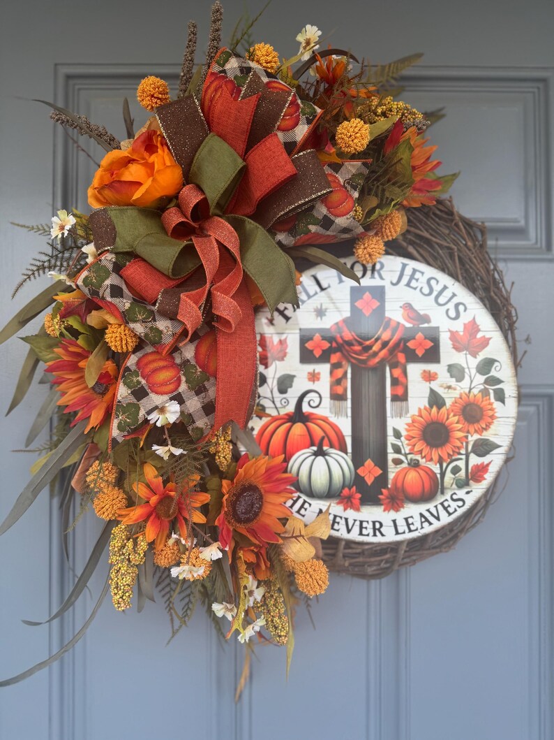 May include: A fall-themed wreath with sunflowers, pumpkins, and a decorative bow. The wreath features a wooden sign with a cross and the text "FALL FOR JESUS HE NEVER LEAVES". The color palette includes orange, brown, and green.