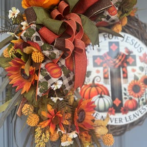 May include: A fall-themed wreath with sunflowers, pumpkins, and decorative ribbons in orange, brown, and green. The wreath includes a circular sign with a cross and the text "FOR JESUS HE NEVER LEAVES".