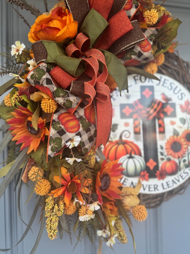 May include: A fall-themed wreath with sunflowers, pumpkins, and decorative ribbons in orange, brown, and green. The wreath includes a circular sign with a cross and the text "FOR JESUS HE NEVER LEAVES".