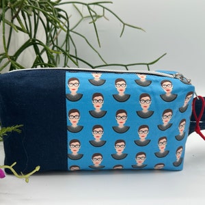 May include: A blue and navy blue zippered pouch with a repeating pattern of a woman's face. The pouch has a red zipper pull and a navy blue strap.