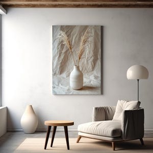 Puede incluir: Una escena interior en tonos neutros. Una impresión en lienzo muestra un jarrón blanco con plantas secas. Una pequeña mesa auxiliar de madera y un sillón crema están en primer plano. Un jarrón blanco grande está en el suelo.