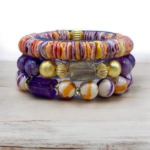 May include: A set of four bracelets featuring a variety of materials and colours. The top bracelet is made of orange, yellow, and purple shell-like pieces. The second bracelet is made of purple and white stones. The third bracelet features a large purple stone, gold beads, and a clear crystal. The bottom bracelet is made of white and orange stones.