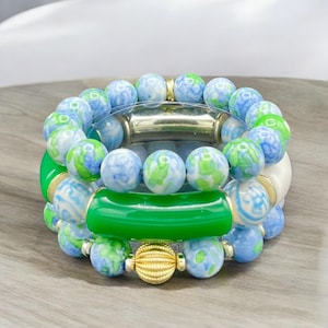 May include: A set of four bracelets featuring blue and green patterned beads. The bracelets are stacked on top of each other, with a green jade bracelet in the middle. The bracelets are accented with gold beads.