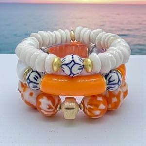 May include: Stacked bracelets featuring white, orange, and blue beads. The bracelets include white shell-like beads, orange cylindrical beads, and round beads with blue and white patterns. Gold-colored accents are visible between the beads. The bracelets are stacked together.