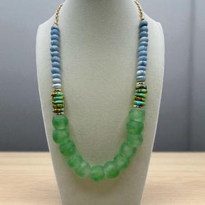 May include: A handmade necklace featuring a variety of beads. The necklace has a gold chain and is composed of light blue, white, green, and gold beads. The focal point is a strand of large, round, light green beads.