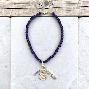 May include: A purple beaded necklace with a gold-colored charm. The charm has the word "AQUARIUS" and a circular pendant with a zodiac symbol. The necklace is on a white and gray surface.