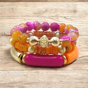 May include: A set of four bracelets featuring a mix of pink, orange, and gold beads. The bracelets are stacked on top of each other, creating a layered look.