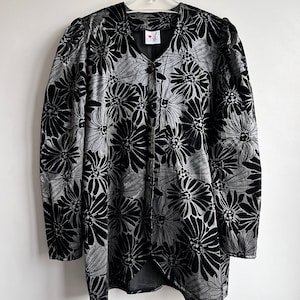 Könnte beinhalten: Schwarze und silberne Jacke mit floralem Muster und Knopfleiste. Die Jacke hat ein großes Gänseblümchenmuster und ein kleines Etikett mit Text. Die Jacke ist aus einem weichen, samtigen Material gefertigt.