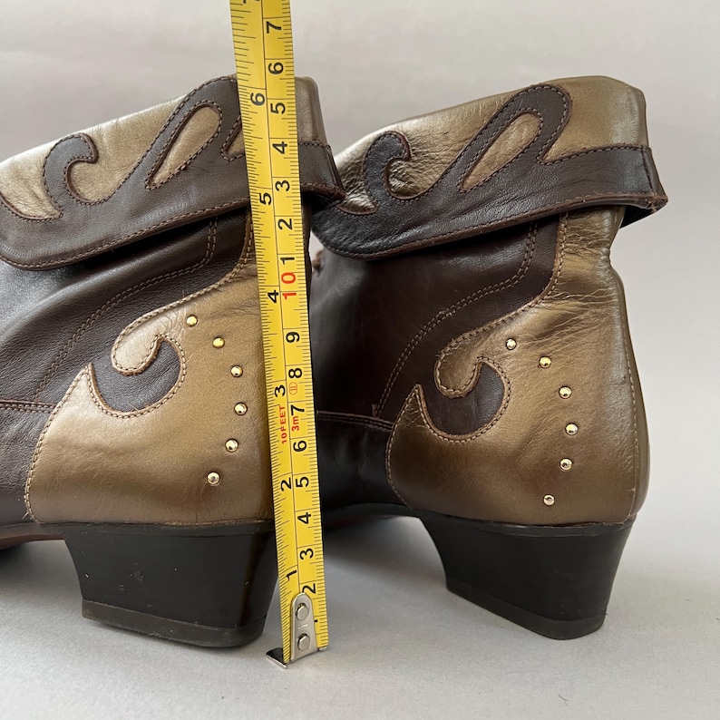 May include: A pair of dark brown and metallic gold leather ankle boots with a fold-over cuff, decorative scroll-shaped overlays, and small gold studs. A yellow measuring tape is positioned vertically, showing a height of approximately 18 cm.