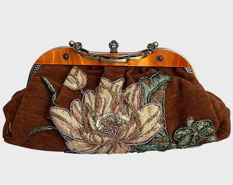 May include: A brown fabric clutch bag featuring an embroidered floral design with gold and green beads. The bag has an amber-colored resin frame and a decorative antique brass-toned metal handle with a central clasp mechanism at the top.