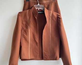 Op de afbeelding: Een bruin blazer met gouden bies en een bijpassende rok. De blazer heeft een mandarijnkraag en lange mouwen. De rok is A-lijn en zit op de taille. Het label op de blazer luidt "Paris Fernand".