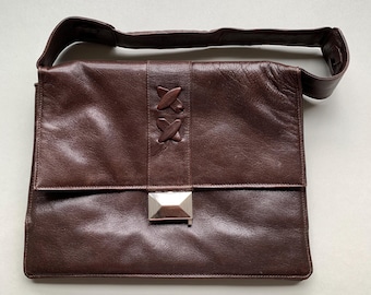 May include: A brown leather shoulder bag with a flap closure and a silver-coloured metal clasp. The bag features decorative stitched details and a matching shoulder strap. The bag is rectangular in shape.