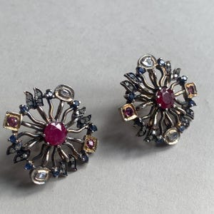 Op de afbeelding: Een paar sierlijke oorbellen met een centrale robijn omgeven door stralende zilverkleurige metalen details. Accentuerende kleine blauwe en goudkleurige vierkante stenen, en zwarte en heldere edelstenen. De oorbellen hebben een bloemen- of sterrenontwerp.