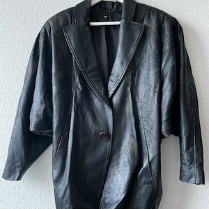 May include: A black leather blazer with a notched collar and a single button closure. The blazer features a floral pattern on the shoulders and sleeves. The jacket is hanging on a white hanger.
