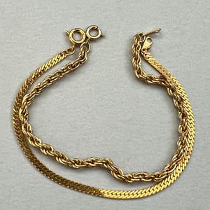 May include: A gold-toned double-strand bracelet. One strand features a herringbone design, while the other has a rope chain style. The bracelet has a lobster clasp closure. The jewelry is displayed on a neutral background.