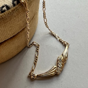 Peut inclure: Un collier doré avec une chaîne délicate et une pièce maîtresse décorative. La pièce maîtresse présente un design incurvé avec trois pierres claires et étincelantes. Le collier est présenté sur une surface neutre.