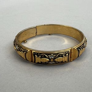 Könnte beinhalten: Ein goldfarbener Klapparmreif mit einem sich wiederholenden Muster aus schwarzen Emaille-Einsätzen und geprägten geometrischen sowie floralen Motiven. Das Armband verfügt über einen sicheren Verschluss und eine glatte, polierte Innenseite.