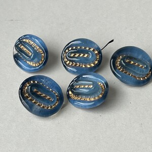 Puede incluir: Cinco botones ovalados azules con un diseño dorado en forma de óvalo en el centro. Los botones tienen un acabado brillante y están dispuestos sobre una superficie gris claro. Los botones son probablemente para coser o hacer manualidades.