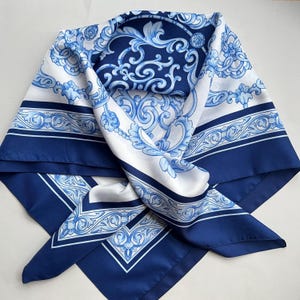 Puede incluir: Pañuelo cuadrado de seda en azul marino y blanco. El pañuelo presenta un diseño barroco detallado con motivos florales y de volutas. El borde es azul marino liso. El pañuelo está doblado para mostrar su patrón.